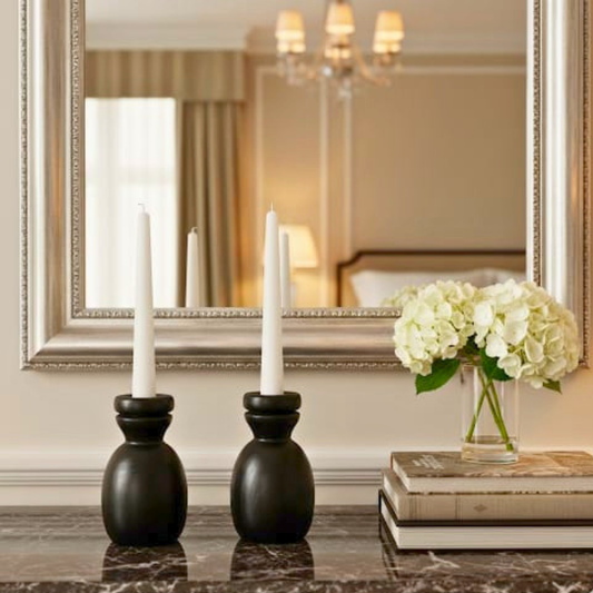 Decorative setup with black candle holders, white candles, and a vase of white flowers in front of a large mirror.