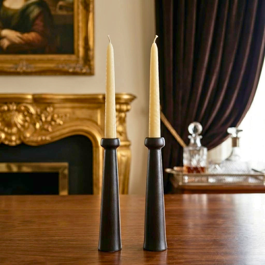 Two tall black candle holders with white candles on a wooden table, with a decorative painting and curtain in the background.