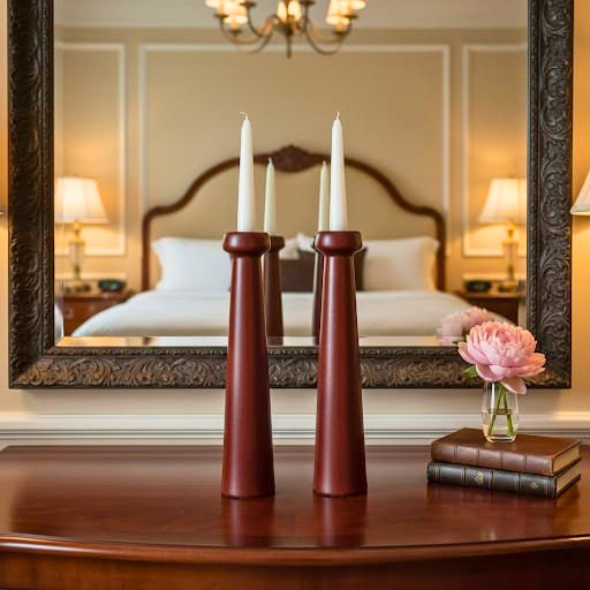 Decorative setup with candles, books, and a flower on a table in a room with a mirror reflecting a bed.